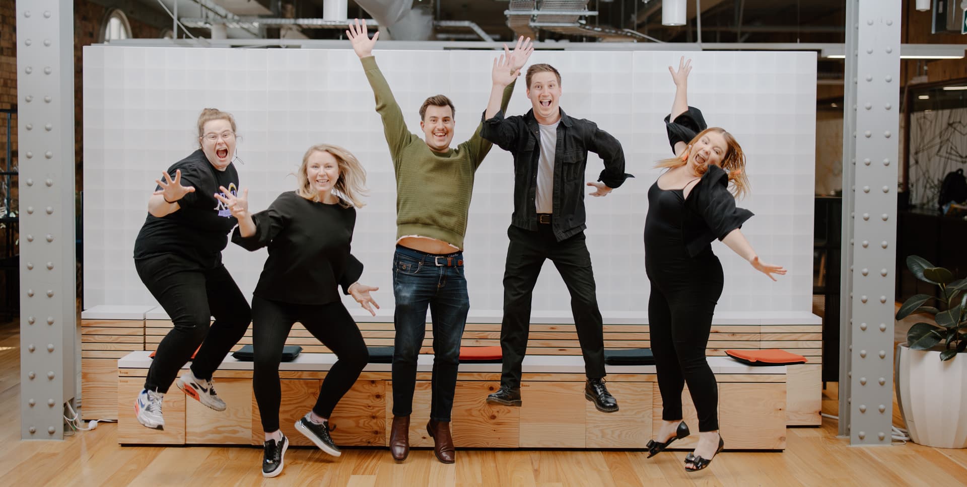 Five people from a SheCodes Brisbane cohort caught mid-jump, arms up and laughing, in a brick-and-timber workspace.